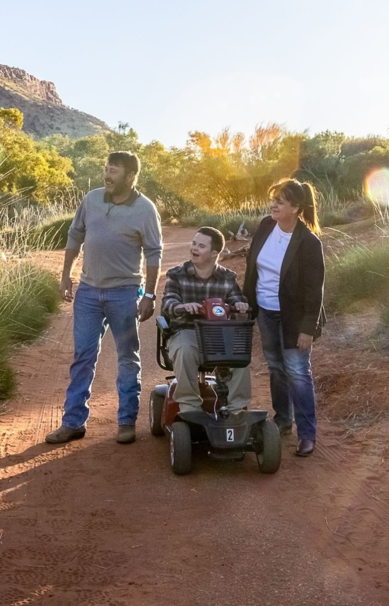 Seorang Pria duduk di skuter elektronik di samping pria lain dan seorang wanita tengah berjalan di antara tanaman hijau dan tanah merah di Alice Springs Desert Park di Alice Springs, Northern Territory Seorang Pria duduk di skuter elektronik di samping pria lain dan seorang wanita tengah berjalan di antara tanaman hijau dan tanah merah di Alice Springs Desert Park di Alice Springs, Northern Territory